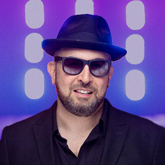 Man wearing a black suit, blue hat, and sunglasses against a purple background with light patterns.